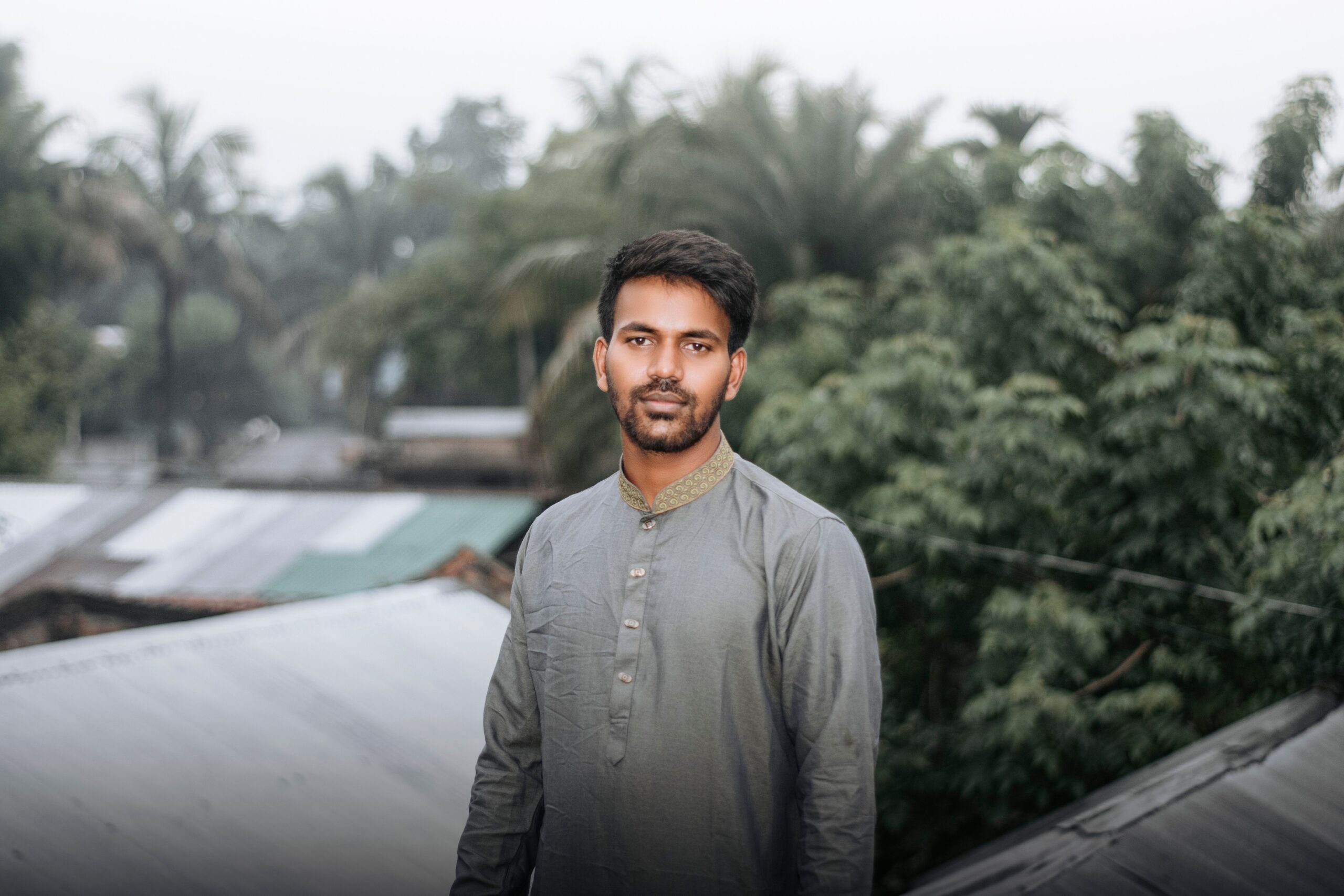 A young South Asian man stands outdoors with lush greenery in the background, exuding calmness.